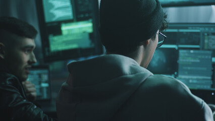 Young male hacker in glasses looking at screen and talking with associate while performing cyber attack on computer in dim room of criminal base