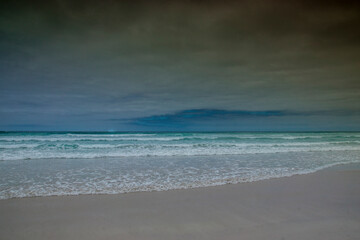 Turquoise, blue and green sea waves on a white sand beach under a grey sky; Tortuga Bay, Galapagos.