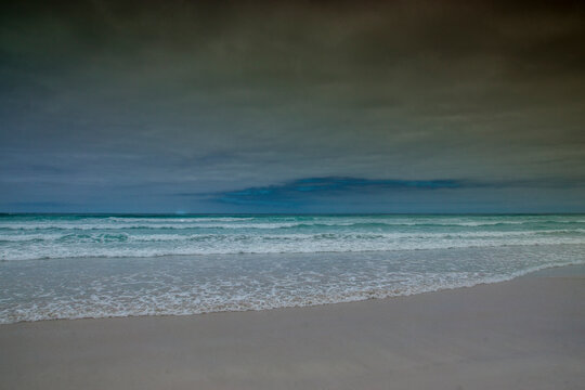 Turquoise, blue and green sea waves on a white sand beach under a grey sky; Tortuga Bay, Galapagos.