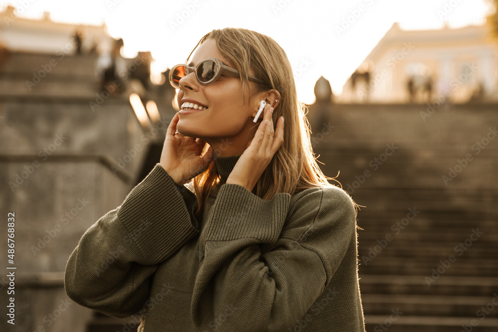 Wall mural Photo beautiful young caucasian woman listening to music, enjoying audio player. Dressed casual clothes, uses wireless headphones, posing on blurred background outside. Fashion, technology concept. - Wall murals