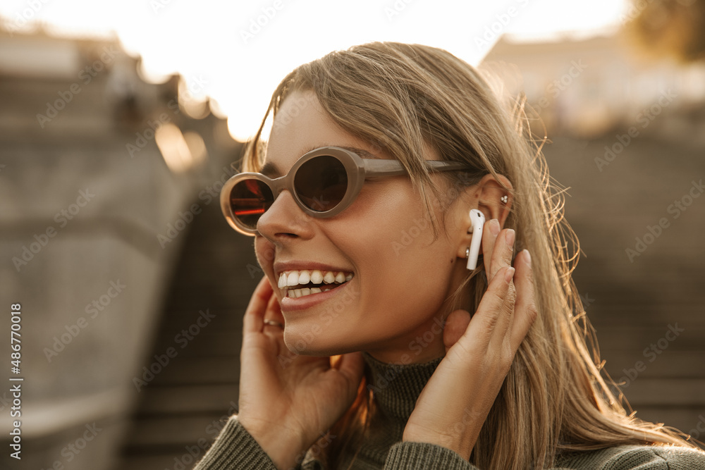 Wall mural Close-up of cute young Hispanic woman in sunglasses holding headphones in her ears. Blonde with snow-white smile on blurred background of city. Beauty and youth concept