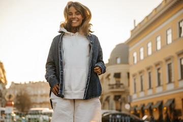 Funny fair-skinned young girl jumps on spot, smiling broadly to her teeth in city center. Tanned blonde with tousled hair is wearing white tracksuit and denim jacket. Holiday life concept