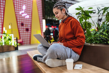 Young woman using her laptop sitting on bench with her legs crossed in a shopping mall