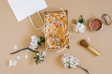 Nail polish and eau de toilette in a gift box with paper shavings, blush and blush brush, apple blossom and white paper bag on a beige background. A gift for a woman for her birthday or mother's day.