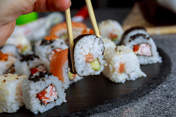 A woman takes sushi with chopsticks from a black plate.