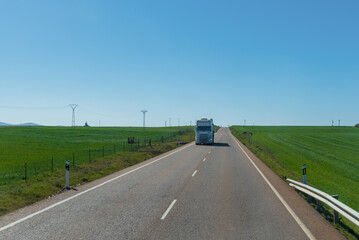 Truck driving along a long straight between green fields on a clear day.