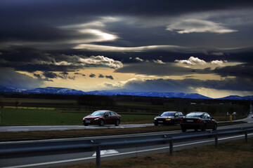 Samochody na drodze szybkiego ruchu, krajobraz wiejski przed zachodem słońca, góry i ciemne chmury.	 - obrazy, fototapety, plakaty