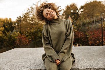Funny young caucasian woman with closed eyes waving her hair sitting on concrete in afternoon....
