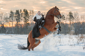 A girl in a white cloak rides a brown horse in winter.