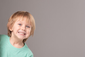 Banner with cheerful child peeking out on edge of white poster, copy space. Portrait of cute little boy looking at the camera on gray studio background. Childhood, health, dentistry, learning concept.