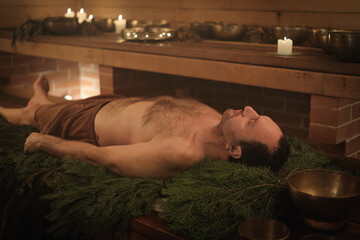 A man lies on spruce branches in a sauna, he is being performed a Tibetan ritual.