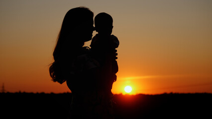 Caring mother holds the baby in her arms and kisses him, unrecognizable silhouette at sunset,...