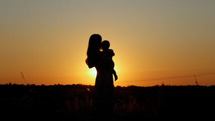 Caring mother holds the baby in her arms and kisses him, unrecognizable silhouette at sunset,...