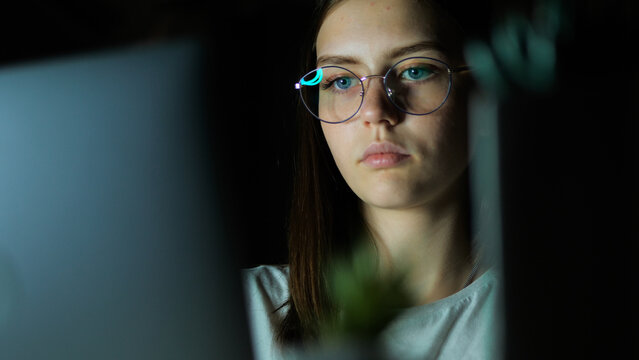 Teenage girl with glasses looks at the monitor, plays a game, reads, studies late at night