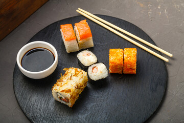 Rolls with fish and shrimp on a round board next to sticks, soy sauce on a dark background.