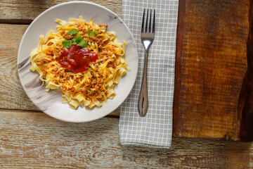 Pasta bolognese garnished with herbs and cheese in a plate on a wooden table on a stand next to a fork and a napkin.
