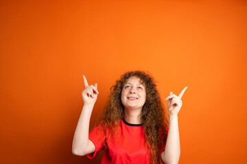 Fan football woman in red t-shirt team uniform looking up and keeping two index fingers up posing on orange background.