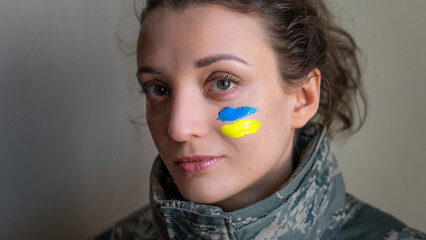 Indoor portrait of young girl with blue and yellow ukrainian flag on her cheek wearing military uniform, mandatory conscription in Ukraine, equality concepts