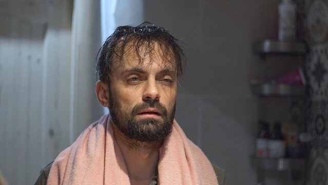 A man in the bathroom in front of a mirror sleepily looks at his reflection with a towel around his neck