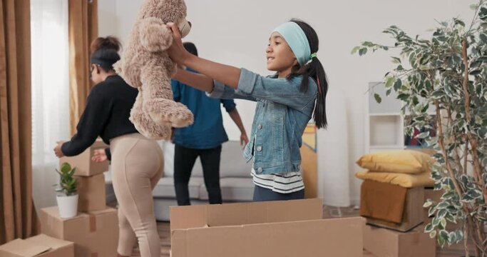 Girl helps parents unpack boxes after moving, finds cardboard box with her toys, pulls out teddy bear, is happy hugs it, mom and dad smile looking at child