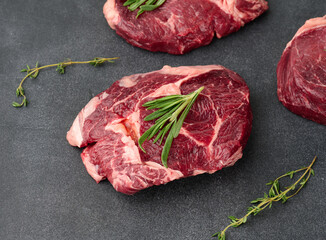 raw piece of beef ribeye with rosemary, thyme on a black table, top view