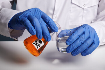 Doctor pours orange poison from flask - labs photo. Scientist pours fake orange juice into a glass .Medic hands in blue nitrile gloves over white desk surface. Toxic fake orange colored drink.