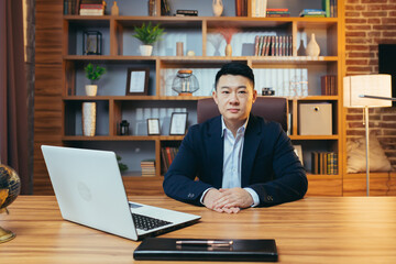 Portrait of a successful insurance agent, an experienced Asian man, sitting at a desk, looking at the camera, in the evening, successful and focused