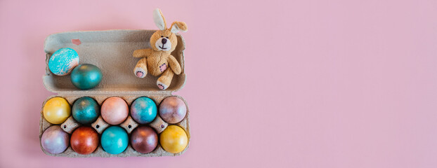 Happy Easter holiday! Fluffy Easter bunny toy sits in the box with multi-colored painted eggs on pink background. Easter egg coloring at home. Preparing for the celebration of Easter. Banner