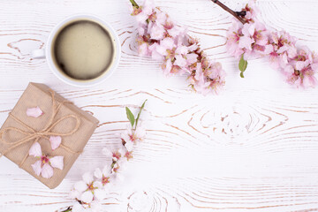 Spring composition with coffee, a gift wrapped in craft paper, spring blossoming almond branches with flowers on a white shabby wooden table. Flat lay, copy space