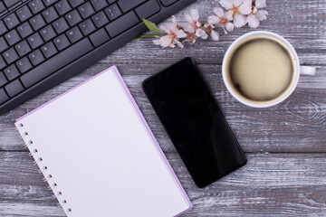 Workspace with keyboard, coffee cup, flower, blank notepad with spring for idea, note, goal, plan writing concept, smart phone on gray whitewashed wooden background. Coffee break, copy space, flat lay