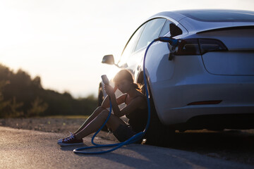 Beautiful young sad girl next to an electric car. Holding a charging cable. Sunset backlight.