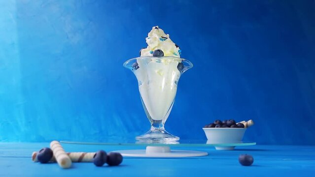 Ice cream with blueberries in a glass rotation on a tray on a wooden table on a blue background. Creative slow motion 4k loop video with speed ramp effect. 