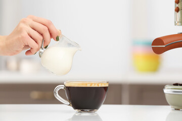 Woman pouring milk into cup of fresh coffee on table in kitchen