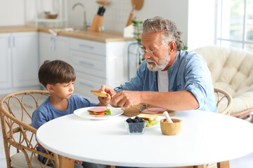 Little boy with his grandfather making sandwich at table in kitchen