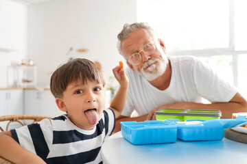 Little boy with his grandfather having fun while packing lunch boxes at table in kitchen