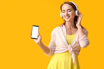 Young redhead woman in headphones with mobile phone on yellow background