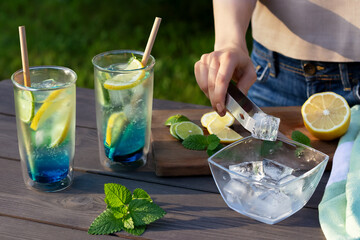 Girl makes summer refreshing cocktails blue lagoon. Close-up of adding ice cubes to glasses....
