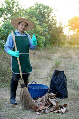 Asian man hold broom, thumb up, stands beside basket of dry leaves, black plastic bag of garbage in forest. Concept : Environment conservation. Collecting dry leaves to make compost                 