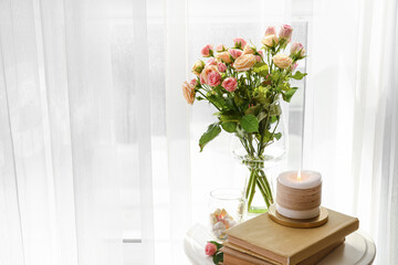 Vase with roses, books and burning candle on table in light room