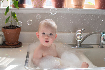 Baby taking bath in the kitchen sink. Child playing with foam and soap bubbles in the sunny bathroom with window. Water fun for kids. Hygiene and skincare for children.
