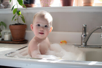 Baby taking bath in the kitchen sink. Child playing with foam and soap bubbles in the sunny bathroom with window. Water fun for kids. Hygiene and skincare for children.