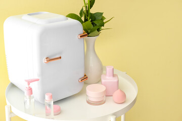 Small refrigerator and cosmetic products on table near color wall