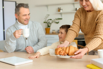 Senior woman putting plate with croissants onto table to her husband and little grandson in kitchen