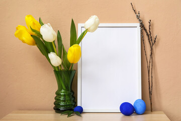 Composition with photo frame, Easter eggs and flowers on table near beige wall