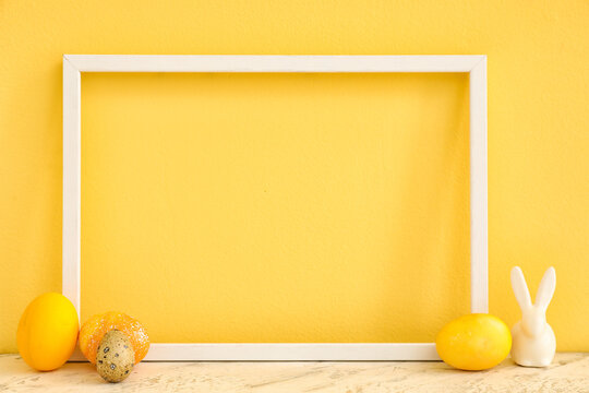 Empty picture frame, Easter eggs and decorative rabbit on table against color background, closeup