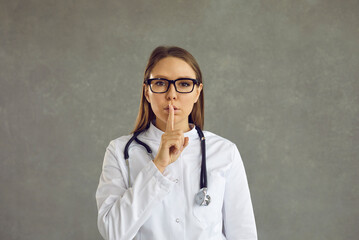 Female doctor in uniform with stethoscope showing shh gesture with index finger holding on lips ask...