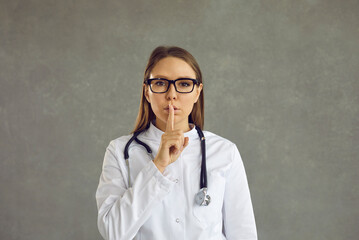 Female doctor in uniform with stethoscope showing shh gesture with index finger holding on lips ask to be quiet studio shot on grey background. Silence and secret, confidentiality concept