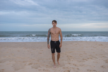 Full height portrait of handsome fit bare-chested young man standing on the beach. guy with tattoo walking by the sea. High quality photo