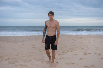 Full height portrait of handsome fit bare-chested young man standing on the beach. guy with tattoo walking by the sea. High quality photo