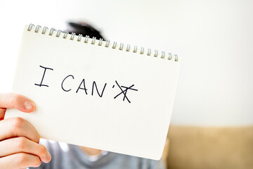 Person’s hand showing book with written word I CAN’T which is crossing lines on letter T, depth of field. Positive attitude and confident mindset to achieve a goal. I CAN self-motivation conceptPerson